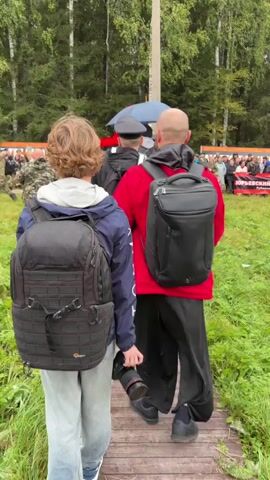 The video opens with a close-up view of three people walking away from the camera on a wooden path. Two of them are wearing backpacks. The person in the center is wearing a red shirt and black pants and is holding a camera. The background shows a line of people standing behind a banner, with trees and a cloudy sky above. The camera then pans to a drone flying in the air, followed by a shot of a forest and a sign that reads "ИНТЕРВЕНЦИЯ (1918-1920)". The scene appears to be an outdoor event or gathering in a wooded area.