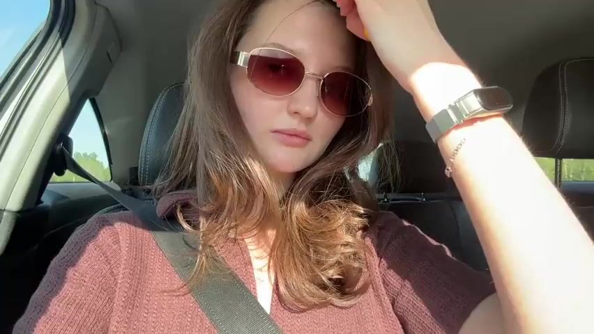 A woman wearing sunglasses and a maroon shirt is sitting in the driver's seat of a car. She is wearing a seatbelt and has a silver watch on her left wrist. She is holding a yellow tube in her right hand and is touching her hair. The car is moving, and trees are visible through the windows. The car's interior is gray and black.