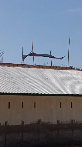 The video shows a side view of a building under construction. The building has a white corrugated metal roof, a light-colored facade with small, evenly spaced rectangular windows, and a dark, rough base. A black mesh tarp is stretched between two wooden poles on top of the roof, with additional wooden poles supporting the structure. The sky is clear and blue. There are no people visible in the frame.