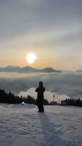 The video shows a person snowboarding on a snowy mountain with a stunning view of the sun setting over a sea of clouds. The person is dressed in snowboarding gear and is standing still, enjoying the scenery. The background includes the sun, mountains, clouds, snow, trees, and snow park features. The scene is peaceful and serene.