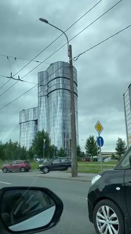 The video shows a large, modern, multi-story building with a glass facade, reflecting the overcast sky. The building has a unique, undulating shape, appearing to be composed of several curved sections. Trees and green spaces are visible around the base of the building. In the foreground, a street is visible with several cars. One car is parked on the side of the road, and other cars are driving on the street. Overhead, power lines and a utility pole are present. The overall environment suggests an urban or suburban setting on a cloudy day. The footage appears to be captured from inside a moving vehicle, as indicated by the reflection of a side-view mirror in the lower left corner of the first few frames.