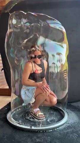A woman wearing sunglasses, a black tank top, and denim shorts squats inside a giant bubble. The bubble is iridescent and reflects a rainbow of colors. She is standing on a circular base with a wet surface. Behind her, a black backdrop is visible. In the final frames, a hand enters the frame from the left, seemingly popping the bubble.