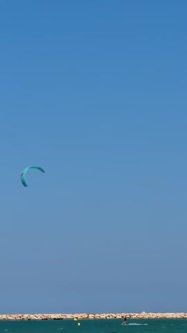 A kite surfer is visible on the right side of the frame, moving from right to left. They are on the ocean, with a breakwater made of rocks forming a line across the lower portion of the image. Yellow buoys are also present in the water near the breakwater. The sky is a clear, vibrant blue. A turquoise kite is visible in the upper left quadrant of the frame, also moving from right to left.
