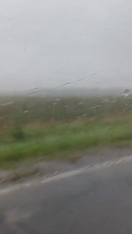 The video shows a blurred, out-of-focus view of a landscape from inside a vehicle, likely a car, moving along a road. The road surface is gray asphalt with yellow lines. To the right of the road, there is a green grassy field that extends into the distance. The sky is overcast and gray, suggesting it might be raining or foggy, as evidenced by streaks on what appears to be the windshield. The overall scene is muted and indistinct due to the motion blur and poor focus.