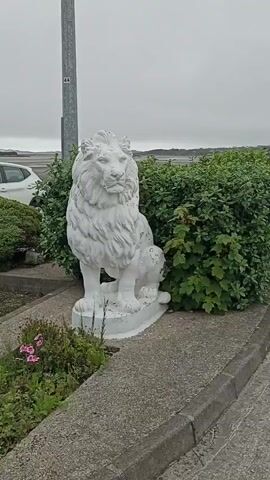 A white lion statue sits in front of a hedge. To the right, there are picnic tables and umbrellas. In the distance, there is a body of water, hills, and buildings. The sky is overcast. A car is partially visible on the left.