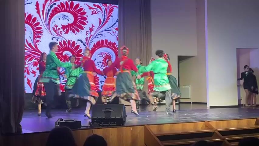 On a stage, a group of children perform a traditional folk dance. There are seven girls and three boys, each dressed in brightly colored folk costumes: red blouses, green shirts, and long skirts with colorful aprons for the girls. The boys wear loose-fitting pants. They dance in a circle, holding hands and stepping lively to the music. Behind them is a large screen displaying a decorative red and white floral pattern. The stage is wooden and extends towards an audience, where a few people are visible in the foreground. A speaker is positioned on the front edge of the stage. To the right of the stage, a modern doorway is visible, and a woman in a black dress stands to the side. The stage is lit with soft, even lighting.