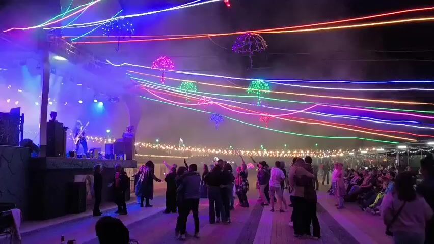 The video captures an outdoor nighttime event with a stage and a crowd. A band performs on a brightly lit stage, with blue and purple spotlights illuminating the performers. Above the stage and the crowd, a canopy of colorful neon lights stretches across the scene, creating a vibrant and festive atmosphere. Several decorated umbrellas are suspended from the lights, adding to the visual spectacle. In the foreground, a group of people are gathered, some dancing and interacting, while others stand and observe the performance. Further back, more people are visible, with some seated and others standing. The ground is paved, and the overall setting appears to be a recreational area or venue designed for entertainment. The air is filled with a light haze, likely from smoke machines or atmospheric effects used during the performance. The lighting and decorations suggest a lively and celebratory occasion.