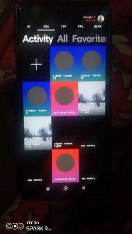 A person is holding a black smartphone with a black screen showing an app interface. The app has a dark theme with white text labels like "Activity", "All", "Favorites", "Staker Redeem", "Creator Redeem", "Collected Daily", "Staked", "Rev Boost", and "New Memory."  The app displays several tiles with obscured content. The person's finger interacts with the screen, tapping or swiping through the tiles. The phone's interface shows a user profile at the top right. The environment appears to be indoors on a surface covered with a red patterned fabric. A "Tecno Spark 9 Pro" logo is visible at the bottom left of the phone. There is indistinct audio in the background.