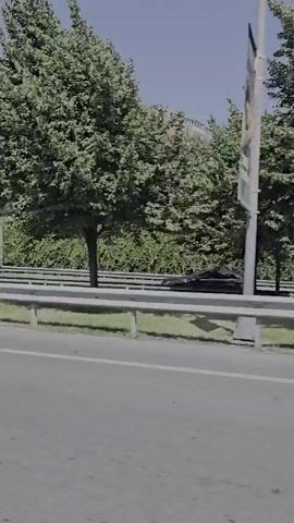 A light-colored car is driving on a road. There are buildings and trees in the background. The car is in the foreground and is the main focus of the image. The sky is clear and blue. There is a truck driving in the distance behind the car.