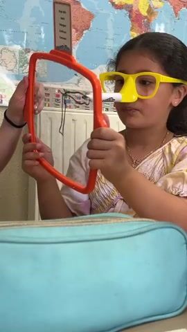 A young girl wearing novelty yellow glasses with a fake nose and a marker attached to the nose is holding an orange object. She is seated at a desk with a blue bag in front of her. Behind her, a world map is displayed on the wall, and a radiator is visible. The girl appears to be interacting with the orange object, possibly using it as a tool for writing or drawing. The marker attached to her glasses is pointed downwards, suggesting it may be used for that purpose. She is looking towards the orange object with a focused expression.