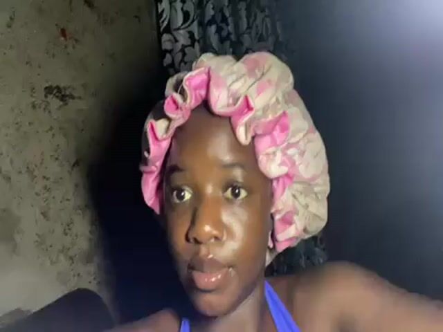 A young woman with dark skin and a pink and white patterned hair bonnet is filming herself in a poorly lit room. She is wearing a blue top. A hand holding a phone flashes light on her face, momentarily illuminating her features. The room appears dark with a wall visible behind her. A black and white patterned curtain can be seen in the background.  Her eyes widen slightly as the light shines on her face.