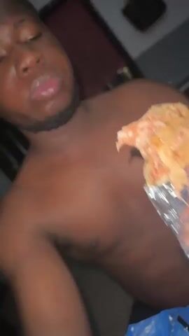 A shirtless Black man with a short beard takes a bite out of a food item wrapped in aluminum foil. He chews and looks at the camera. A blue plastic bag is visible below him. A purse hangs on the wall in the background.