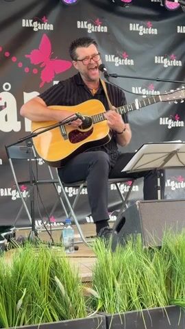 A man with glasses and a black collared shirt is sitting on a folding chair and playing an acoustic guitar and singing. He is performing on a stage with a black backdrop that has the word "BAKGARDEN" repeated multiple times, as well as a pink butterfly graphic and some pink dots. There are stage lights above him. In front of him on the stage is a music stand with a piece of paper and a black speaker. To his left is a bottle of water on the floor. In the foreground, there is tall green grass.