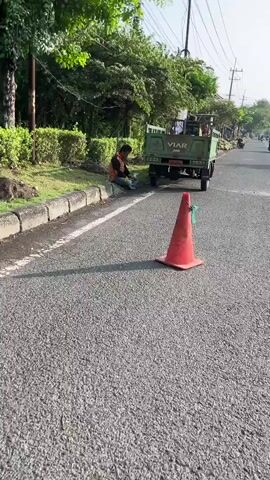 The video opens with a bright, sunny outdoor scene. In the foreground, a red traffic cone with green string attached to the top sits on an asphalt road. Beyond the cone, a green truck with the brand name "VIAR" visible on the back is parked on the right side of the road. A person wearing a light-colored shirt and orange fabric is seated on the grass next to the road, facing away from the camera, appearing to be resting or working on something. The truck bed contains some items. The road appears to be a two-lane road with a white line marking the lanes. Lush green trees and bushes line the left side of the road, and a wire pole with electrical lines is visible in the background on the right. As the video progresses, the camera pans slightly to the right. A person wearing a green shirt and an orange head covering is seen standing near the back of the truck. Another person in a green shirt is riding a motorcycle in the background on the right side of the road. The scene suggests a roadside setting, possibly in a rural or semi-urban area, during daylight hours.