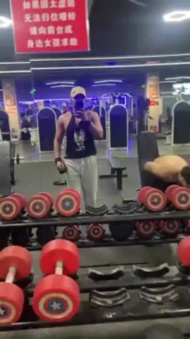 The video shows the reflection in a gym mirror of two young men. One man is facing the mirror, shirtless, seated on a bench, and seems to be putting on a black jacket or sweatshirt. The other man is standing near the mirror, facing it, taking a selfie with his phone. He is wearing a tank top, light-colored sweatpants, and a tan baseball cap. Behind the two men and reflected in the mirror is gym equipment, including weight machines and various weights. There is a red sign with white lettering hanging above the mirror, presumably providing instructions or rules for the gym. In the foreground is a rack of red dumbbells with a Captain America design.