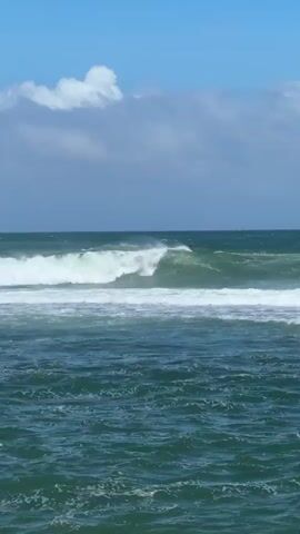 This video presents a dynamic ocean scene in Walgama, Sri Lanka, under a bright, partly cloudy sky. The deep blue-green sea is characterized by powerful waves that continuously crest and break, rolling towards the foreground. Each wave features a vibrant white foam as it breaks, with visible spray being whipped off the top by strong winds, suggesting blustery conditions. The horizon is visible in the distance, separating the active ocean from the light blue sky, which is dotted with large, fluffy white clouds. The ambient sound of the video is dominated by the constant roar of the waves crashing and the distinct rush of the wind. No people, boats, or other man-made structures are visible, allowing for an immersive focus on the natural spectacle of the sea.