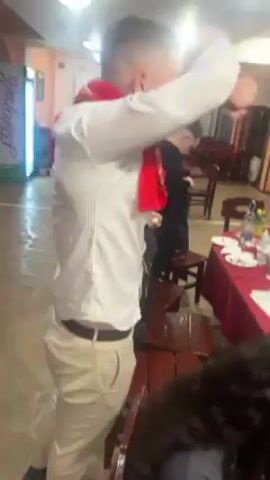 The video shows a group of people at what appears to be a celebratory gathering inside a restaurant. The main focus is on the tables laden with food, drinks, and used dishes, suggesting a meal is either concluding or has recently ended. People are milling about, some gesturing and appearing animated, while others are seated. The environment includes typical restaurant decor with chairs, tables, and doors leading outside. There are decorations such as flags. The overall impression is of a lively, perhaps slightly messy, event.