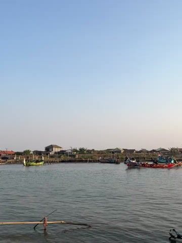 The video shows a serene waterfront scene with a clear blue sky. In the foreground, a wooden structure made of bamboo sticks emerges from the calm water. In the middle ground, several colorful fishing boats are visible on the water. Some of these boats are docked along the shore, while others appear to be in motion. The background features a strip of land with houses and lush vegetation, suggesting a coastal village or settlement. The overall atmosphere is peaceful and tranquil, with the gentle movement of the water and the presence of the fishing boats indicating a typical day in a fishing community.