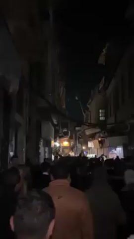 The video shows a densely packed crowd moving through a narrow alleyway at night.  The buildings are close together and tall, with a network of wires crisscrossing overhead.  A few lit signs are visible along the buildings.  The crowd appears to be celebrating or protesting, as there is cheering and shouting.  The camera is held at eye level and moves with the crowd.