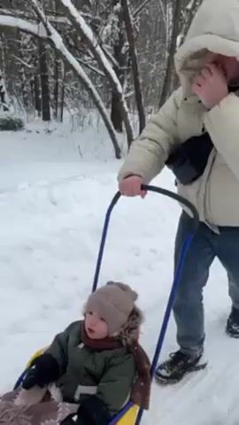 A person is pulling a child on a sled through the snow. The child is bundled up in a hat and jacket and covered with a blanket. The person pulling the sled is wearing a beige puffer jacket and jeans. They are walking on a snowy path through a forest. The trees are covered in snow.