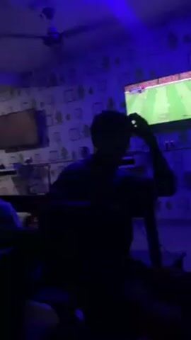 The video depicts a group of individuals watching television in a dimly lit room. The room is furnished with pews and a ceiling fan, and the walls are adorned with patterned wallpaper. Two televisions are visible, one playing what appears to be a sports game. The people in the room are mostly silhouettes, their faces obscured by the low lighting, but they appear to be engaged in watching the screen. The room is illuminated by a combination of the television screens and an overhead blue light, creating a relaxed, casual atmosphere.