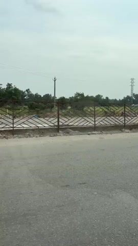A car is driving along a road, with a side view mirror visible in the foreground. The mirror reflects the inside of the car, showing a person holding a phone and possibly a cigarette. Outside the car, there is a metal fence with a geometric pattern. Beyond the fence, there is an open field with some trees and vegetation. In the distance, there are power lines and a tower. The sky is overcast. The scene appears to be a roadside view in a rural or semi-rural area.