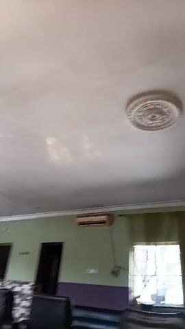 The camera pans across the interior of a room. The ceiling is white with decorative molding. A chandelier is visible. The walls are painted in two tones of green and purple. A window is covered by green curtains. An air conditioning unit is mounted on the wall. Several black chairs are in the room. A large fan is also visible.