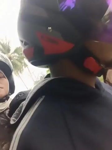 The video begins with a close-up shot of a person wearing a black helmet with a reflective visor and a dark jacket with white stripes. The camera then pans to the right, revealing another person wearing a light brown hijab, also on a motorcycle. The scene appears to be outdoors, with trees and a building visible in the background. The video continues to show the two individuals riding on a road, with other vehicles and palm trees in the distance. The overall atmosphere seems casual, as if documenting a journey or a casual outing.