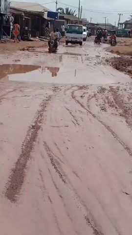 The video shows a muddy road with several vehicles and people. In the foreground, the camera is positioned on a motorcycle, showing the handlebars and the rider's hands. The road is filled with large puddles of water and mud, with tire tracks visible. In the background, there are several buildings lining the road, which appear to be shops or market stalls. People are seen walking and standing around the shops. A white truck is driving down the road, followed by a motorcycle. Another motorcycle is seen further down the road. The sky is overcast. The overall impression is of a busy, albeit poorly maintained, urban or semi-urban environment.