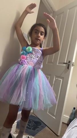 A young girl dressed in a purple and pastel colored tutu dress, with floral embellishments, dances in a doorway. Her arms are raised above her head. The room is dimly lit and appears to be a bedroom. A white door is visible behind her, and a partial view of another doorway is visible in the background.
