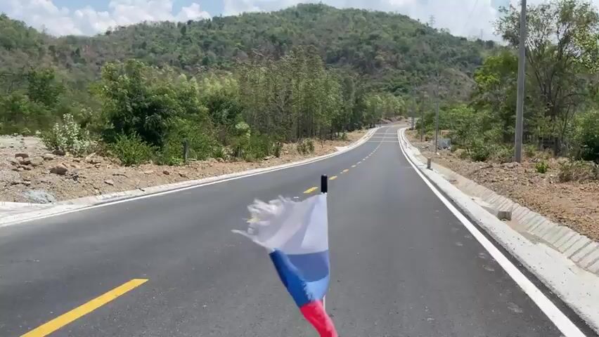 The video shows a new paved road winding through a rural area. The road has yellow lane markings, white edge lines, and concrete barriers along the sides. There are trees and vegetation on both sides of the road, and a small mountain is visible in the background. Power lines run alongside the road. A white flag is visible in the foreground, likely attached to the vehicle filming. The sky is blue with some clouds.