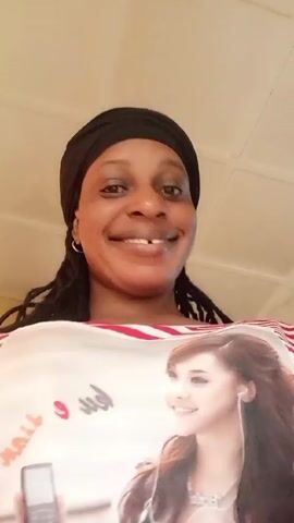 A medium shot features a woman with dark skin, wearing a black head wrap and a white shirt with red stripes and a large image of a younger woman. The woman in the video is smiling, revealing a gap between her front teeth. Her hair appears to be styled in braids or dreadlocks that fall around her shoulders. The camera is positioned slightly below her, looking upwards. The background consists of a plain, light-colored ceiling, with subtle lines suggesting panels. There are sounds of a baby in the background.