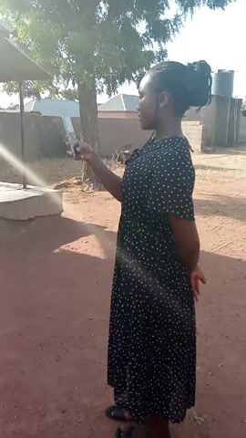 A person in a black, full-length dress with white polka dots stands on reddish-brown ground, holding a phone up in front of their face. The person has dark skin and their hair is pulled up into a bun on top of their head. The person appears to be looking at the phone screen. Behind the person is a tree, a small structure with a roof, and several buildings in the distance. The lighting is bright and sunny.