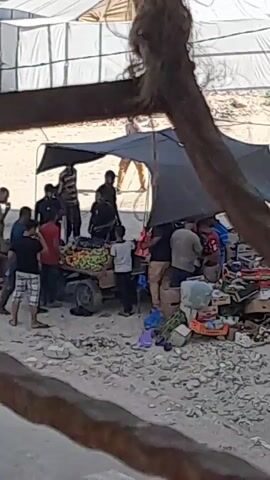 The video shows a group of people gathered around a makeshift stall, possibly a market or an aid distribution point. The stall is covered by a dark tarp, providing shade. Various items, including boxes, bags, and what appear to be food items like bananas, are visible on and around the stall. Several people, including men, women, and children, are interacting with the stall and each other. Some individuals are carrying bags or boxes, and there is a sense of activity and movement within the scene. The ground appears to be covered in gravel or sand, and the background suggests an outdoor environment, possibly a camp or a less developed area, with a large white fabric structure in the distance. The overall impression is of a busy, informal gathering centered around the distribution or sale of goods.