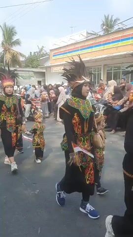 A parade is taking place outdoors on a paved road. The participants are dressed in traditional attire with vibrant, colorful embroidery on black garments, and some wear feathered headdresses. Several individuals are holding small Indonesian flags. A child in traditional dress is walking alongside an adult, holding a flag. A crowd of onlookers lines the street, some on motorcycles. In the background, there are buildings, including a store with an awning. The sky is visible and appears to be clear.