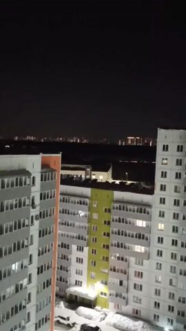 A night view from a high vantage point shows a residential area with multiple tall apartment buildings. The buildings are mainly grey and white with a few sections painted in orange and green. Snow is visible at the base of the buildings and in the surrounding area. In the distance, city lights create a skyline against the dark night sky. The camera is steady, and there are no discernible actions or interactions occurring in the scene. The environment is urban and quiet.
