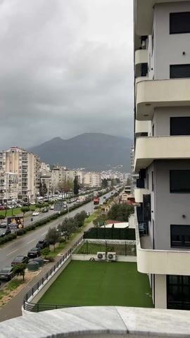 The video captures an elevated view of a city street on an overcast and stormy day, looking towards a dark mountain range in the distance.

In the immediate foreground, a white balcony railing with a textured surface is visible at the bottom of the frame. To the right, a section of a modern apartment building with white and light grey facades and rounded balconies is shown. Below this building, there is a small artificial grass area enclosed by a dark fence, with two air conditioning units situated next to the fence.

The midground features a wide, multi-lane road running horizontally through the scene. A green median strip, planted with small trees (possibly olive trees) and shrubs, separates the two directions of traffic. Numerous cars are visible, both parked along the sides of the road and driving on it, suggesting moderate traffic. On the left side of the road, several large, multi-story residential buildings, mostly light-colored with multiple balconies, line the street. Many cars are parked in front of these buildings. A large billboard with text in what appears to be a Slavic language is visible on the left, advertising something like "MALA KUHINA" (Small Kitchen).

In the background, more buildings extend along the road into the distance. A prominent, dark mountain range dominates the horizon, its peaks partially obscured by heavy, dark grey clouds that fill almost the entire sky. The general lighting is dull and diffused, indicative of the stormy weather.

The audio includes the sound of wind and car traffic. A female voice explicitly states, "The massive storm is rolling in," confirming the visual impression of an approaching weather event. The overall atmosphere is gloomy due to the dark clouds and impending storm.