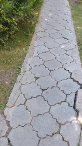 The video shows a first-person perspective of walking down a paved path. The path is made of interlocking concrete pavers with a decorative clover-like shape. To the left of the path is a dense hedge of dark green coniferous trees. To the right of the path is a grassy area. As the person walks further, the path curves slightly to the right, and in the distance, a white building with balconies can be seen between the trees. The lighting suggests it is daytime, likely sunny, with shadows visible on the path.