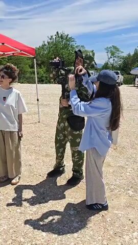The video shows a man and a woman in an outdoor setting. In the first shot, a woman wearing a blue shirt and a baseball cap is helping a man in camouflage attire adjust a camera. The man is holding the camera on a stand. In the second shot, the man in camouflage is replaced by a man wearing a green and yellow athletic shirt, black shorts, and sunglasses, with a medal around his neck. The woman, now wearing a white t-shirt, is holding a small object near his chest. Both men are facing the woman. In the background, there is a red tent and a blue archway with the word "СТАРТ" written on it. Green hills are visible in the distance under a clear sky.