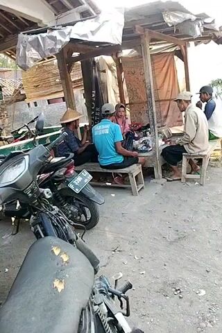 A group of four men and one boy are gathered around a small, open-air shelter constructed from wood and corrugated metal roofing. Two motorcycles are parked in the foreground, one black and the other partially visible. The shelter has a dirt floor and appears to be a roadside rest stop or a small shop. The men are seated on wooden benches and stools. One man, wearing a conical hat, is talking to another man seated next to him. A third man, in a blue t-shirt, is facing away from the camera. The fourth man, wearing a light-colored long-sleeved shirt and a cap, is seated and looking at a book or tablet. A young boy is seated between two of the men. The background shows a simple building with a tiled roof. The overall scene suggests a casual gathering in a rural or semi-rural setting.