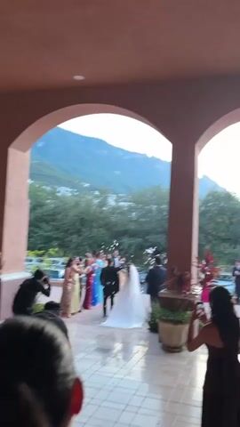 A wedding party is exiting a building. The bride and groom, dressed in wedding attire, are walking away from the camera, and guests are throwing confetti at them. The bride is wearing a white wedding dress with a long veil, and the groom is in a black tuxedo. A group of guests, mostly women in colorful dresses, are standing to the left of the bride and groom, throwing confetti. There are two photographers in the foreground, one on the left and one on the right, capturing the moment. The background shows lush green trees and a mountainous landscape. The scene appears to be an outdoor wedding reception or ceremony exit.
