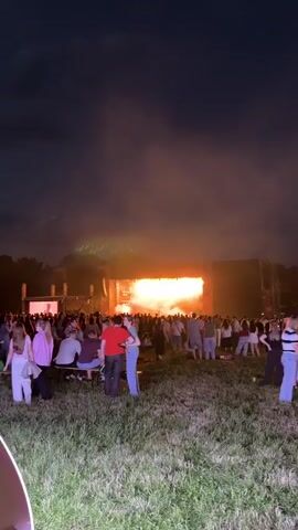 A nighttime outdoor concert scene shows a large crowd gathered on a grassy field facing a brightly lit stage. The stage is adorned with numerous lights and screens displaying visuals, including a large circular image of a man speaking into a microphone. Smoke and pyrotechnics create a dramatic atmosphere on stage, with vibrant orange and yellow lights illuminating the performers and the surrounding haze. The crowd, a mix of adults of various ethnicities, are standing, sitting on the grass, and interacting with each other. Some individuals are holding up phones, possibly recording the event. The overall environment suggests a lively and energetic atmosphere at a music festival or concert.
