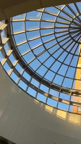 This video shows a low-angle, upward shot of a glass dome ceiling with a radial design. Through the transparent ceiling, a tall yellow building with multiple windows is visible against a bright blue sky. Sunlight casts shadows on the interior of the building, highlighting the architectural elements. The camera appears to be stationary.
