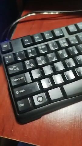 The video shows a close-up, angled shot of a black computer keyboard resting on a dark red, wood-grained surface. The keyboard is oriented diagonally, with the top left corner closer to the camera. The keys are black with white lettering, appearing to be in a Cyrillic alphabet. The lighting is dim, casting shadows and creating reflections on the keys. The video appears to be a continuous, static shot of the keyboard, with no visible movement or activity.