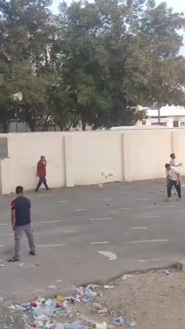 The video shows several children and two adults playing a game in an empty parking lot. The game appears to be some form of stickball. There is garbage scattered on the ground. A large tree looms over a wall in the background. Beyond the wall, a tall, multi-story building is visible. Several of the adults stand to the side, watching the game.