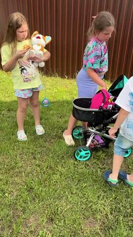 The video shows a summer day outdoors in a grassy area next to a wooden fence. Three children are present. On the left, a girl wearing a yellow shirt with a graphic design and patterned shorts holds a stuffed animal and a toy. She is looking at the other children. In the center, a girl with blonde hair tied back and wearing a pink and green floral crop top and light purple leggings is interacting with a baby stroller. The stroller is black with teal accents and contains a pink blanket. The girl is putting something into the stroller. On the right, a boy with blonde hair, wearing a white short-sleeved shirt with a blue palm tree and beach print and denim shorts, is standing and looking at the camera. He smiles. The camera briefly pans to show another girl in the background, wearing a pink shirt and white shorts, near a table with drinks.