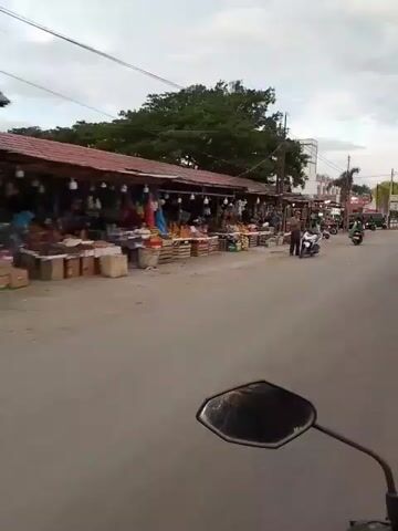 The video shows a street scene with market stalls on the left side of the road and a building labeled "POLISI POLICE" on the right. There are several motorcycles and a few people visible. The market stalls appear to be selling fruits and other goods. The weather is overcast. The video is shot from the perspective of someone riding a motorcycle.