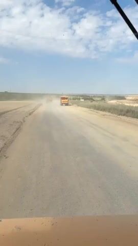 A first-person perspective video captures a dusty, unpaved road on a clear, sunny day with scattered clouds. Two large, orange dump trucks are visible in the distance, driving away from the camera. The truck closest to the camera is in the right lane, kicking up a significant amount of dust as it moves. The road appears to be under construction or in a rural area, with dry, flat terrain on either side. The foreground shows the hood or dashboard of a vehicle, also orange, implying the video is being recorded from another vehicle. The overall impression is one of ongoing work or travel in a dry, arid environment.