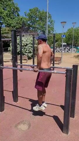 A man is doing dips on parallel bars in a park on a sunny day. He is wearing a blue baseball cap, burgundy shorts, and white athletic shoes. He is shirtless and has tattoos on his arms. The bars are silver and supported by black metal posts. The ground beneath the bars is red and appears to be made of a soft material. In the background, there are trees, benches, light poles, and graffiti on a wall.