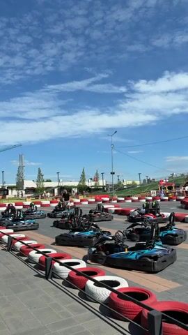 The video shows an outdoor go-kart track on a sunny day with a partly cloudy sky. Several go-karts are parked in rows on the track, with some people walking around them. The track is lined with tires that are painted red and white. In the background, there are buildings, trees, and a crane. One person walks towards the go-karts in the foreground, while others are visible in the background, some near the go-karts and others further away on the track. The go-karts appear to be electric, with blue and black designs. The overall atmosphere suggests a recreational facility ready for use.